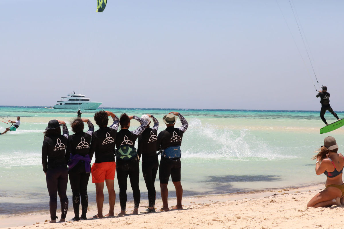 Kite-Tribe_family-on-the-beach group of people on the beach watching a kitesurfer jump. everyone is wearing a kite tribe tshirt