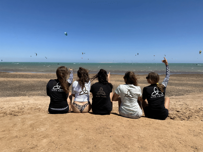 Kite-tribe-el-gouna-team-shirts-beach group of 5 girls sitting on the kite beach in El Gouna in Egypt, looking at the kites in the water. The image shows the girls from the back, all wearing a Kite Tribe T-Shirt
