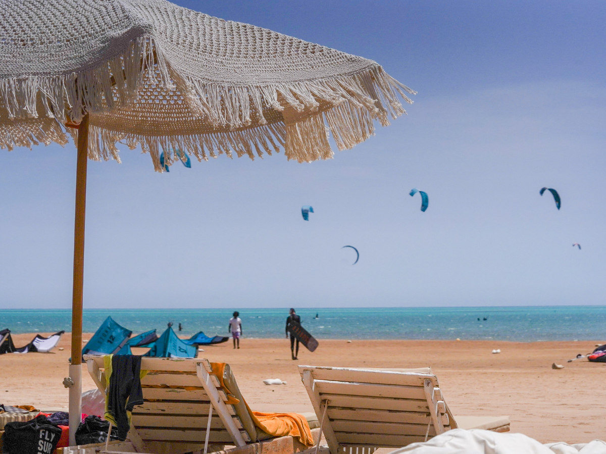 welcometokitetribe group of 5 girls sitting on the kite beach in El Gouna in Egypt, looking at the kites in the water. The image shows the girls from the back, all wearing a Kite Tribe T-Shirt